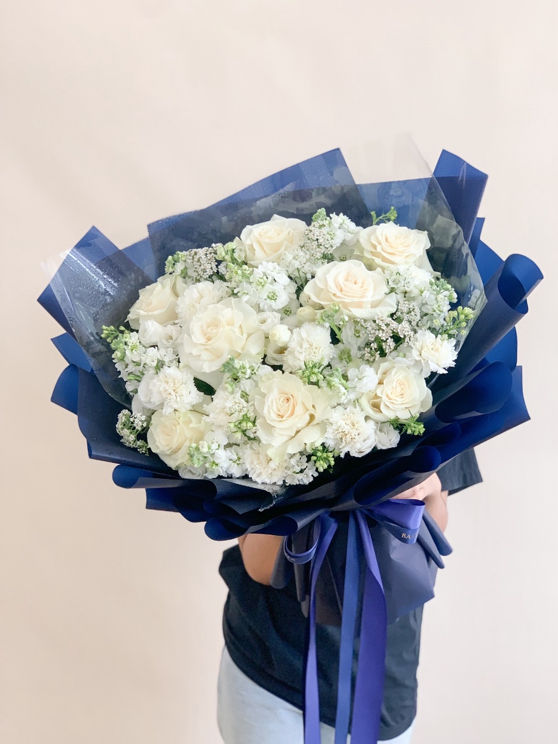 Bouquet of white roses, carnations, and baby’s breath wrapped in luxurious navy blue paper.