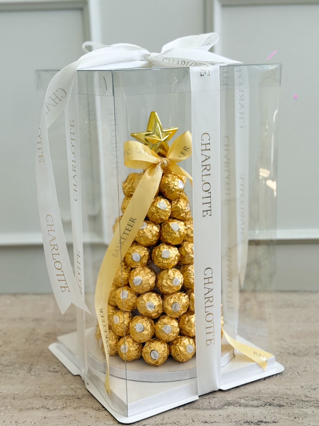 A Golden Ferrero Rocher Christmas Tree, arranged in a pyramid shape and topped with a gold star, is wrapped in yellow and white "CHARLOTTE" ribbons and displayed inside a clear gift box.