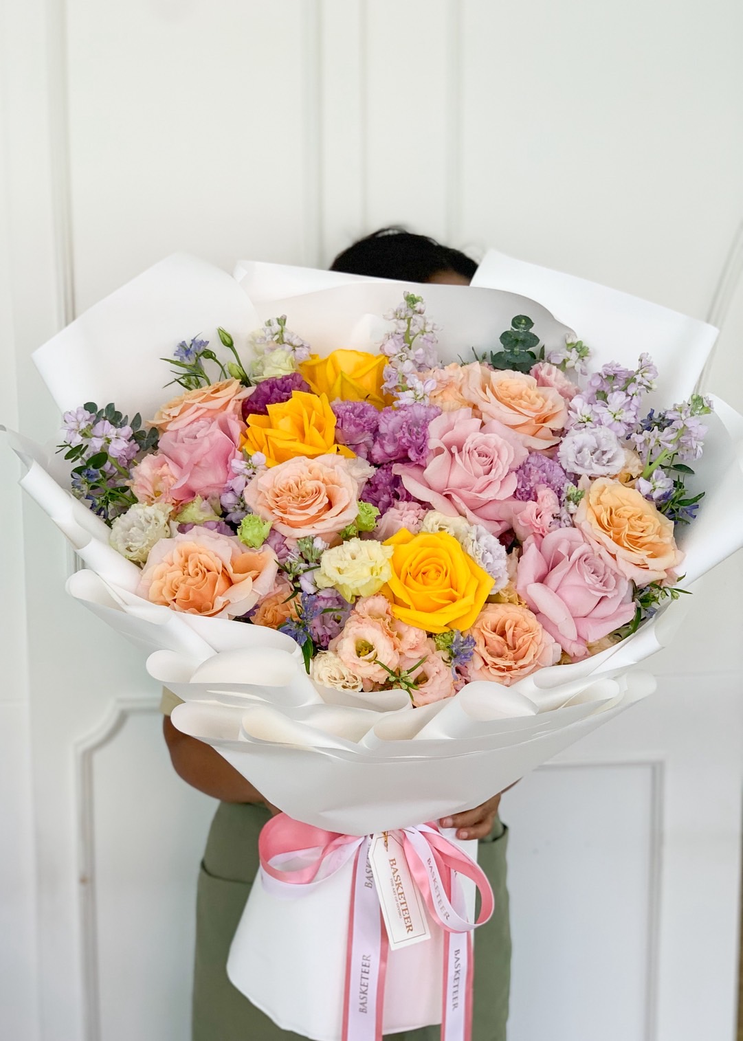 Large bouquet with yellow, peach, pink, and purple roses, wrapped in white paper and pink ribbon