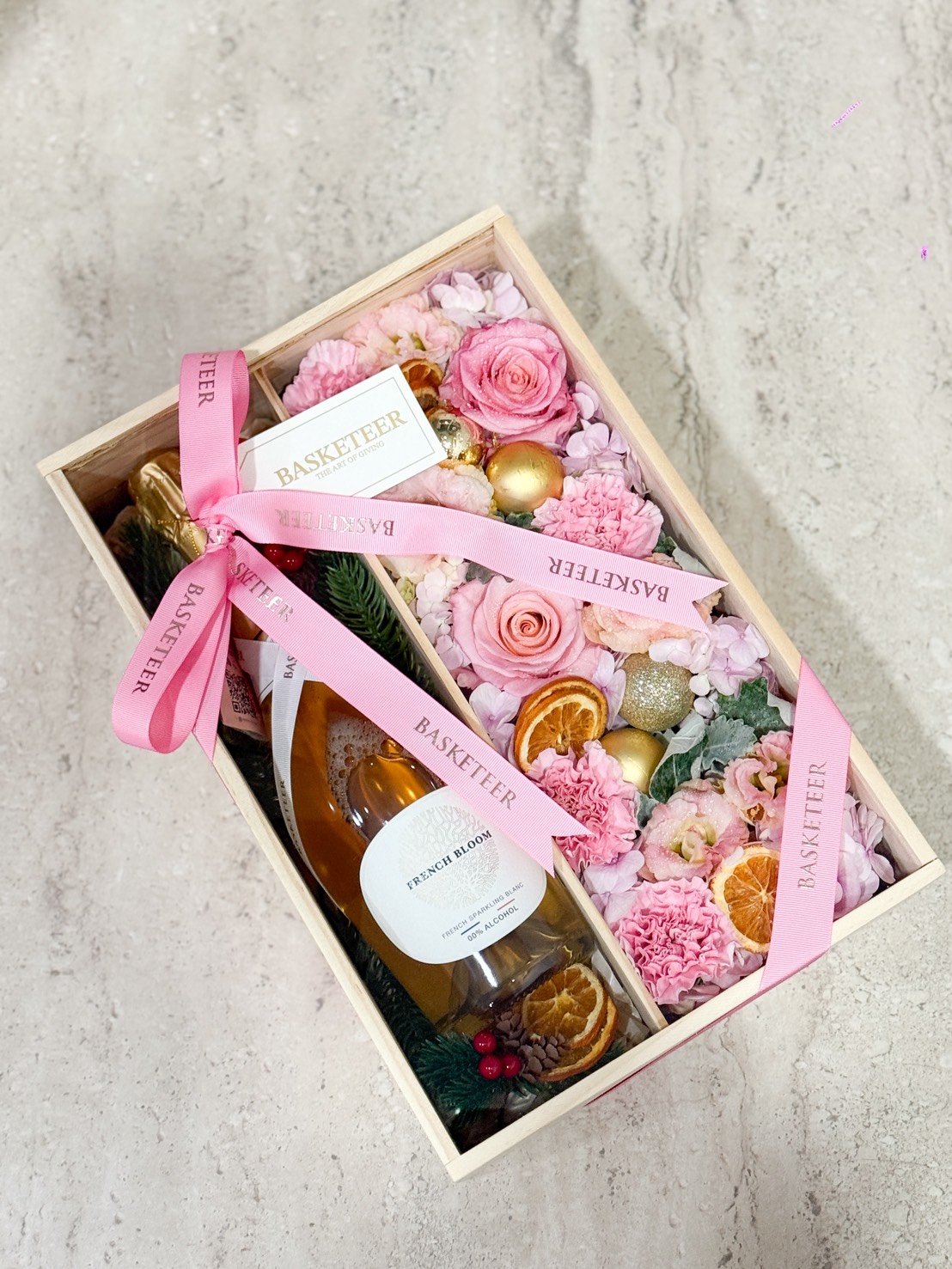 The Blossom Charm Luxury Floral Gift Box features a wooden box with French Bloom, pink flowers, dried orange slices, golden ornaments, and a pink "BASKETEER" ribbon, beautifully arranged on a light-coloured surface.