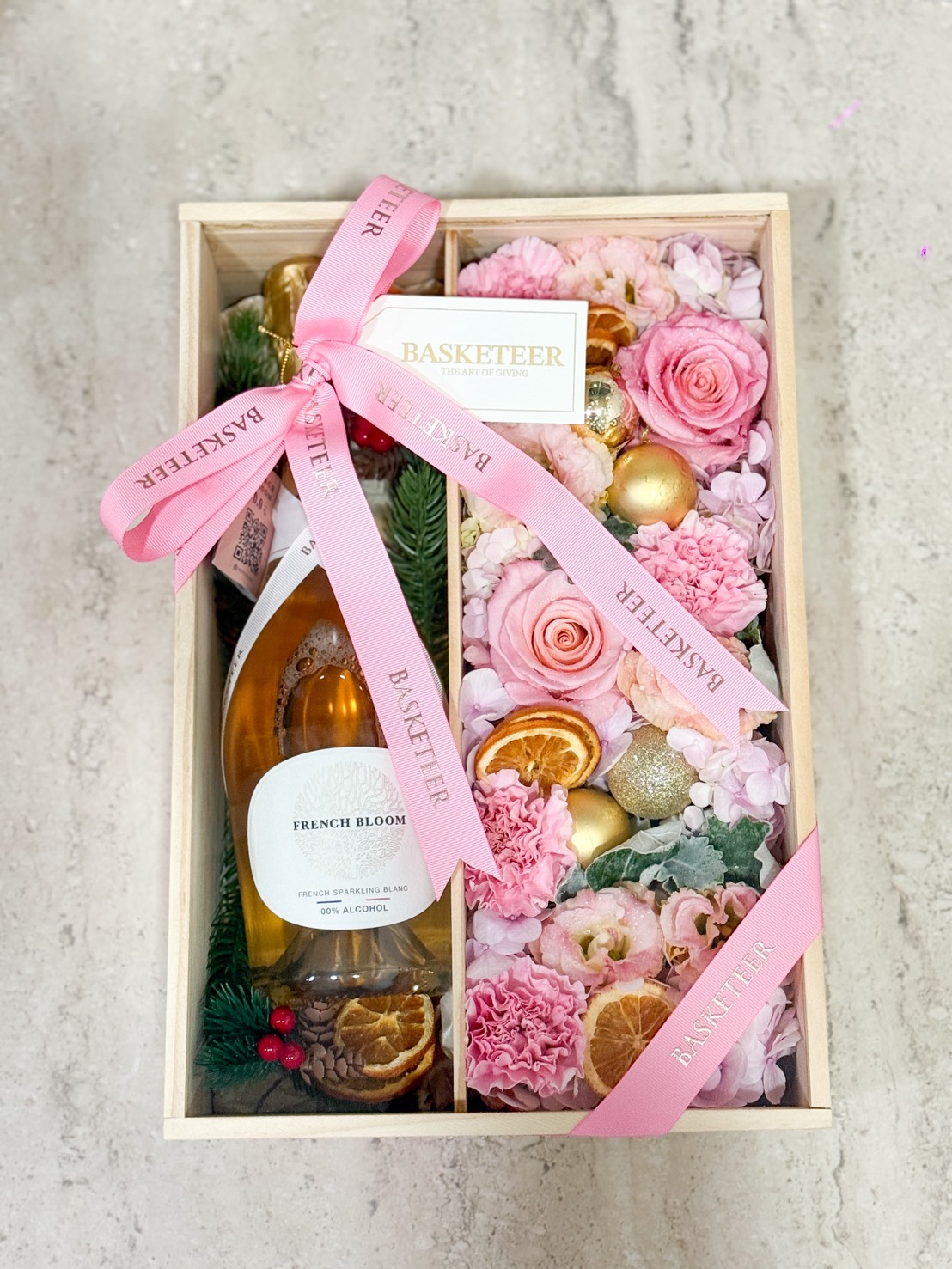 The Blossom Charm Luxury Floral Gift Box features a bottle of French Bloom sparkling wine, pink roses, carnations, dried orange slices, gold ornaments, and lush greenery, all elegantly arranged with "BASKETEER" ribbons on a light marble surface.