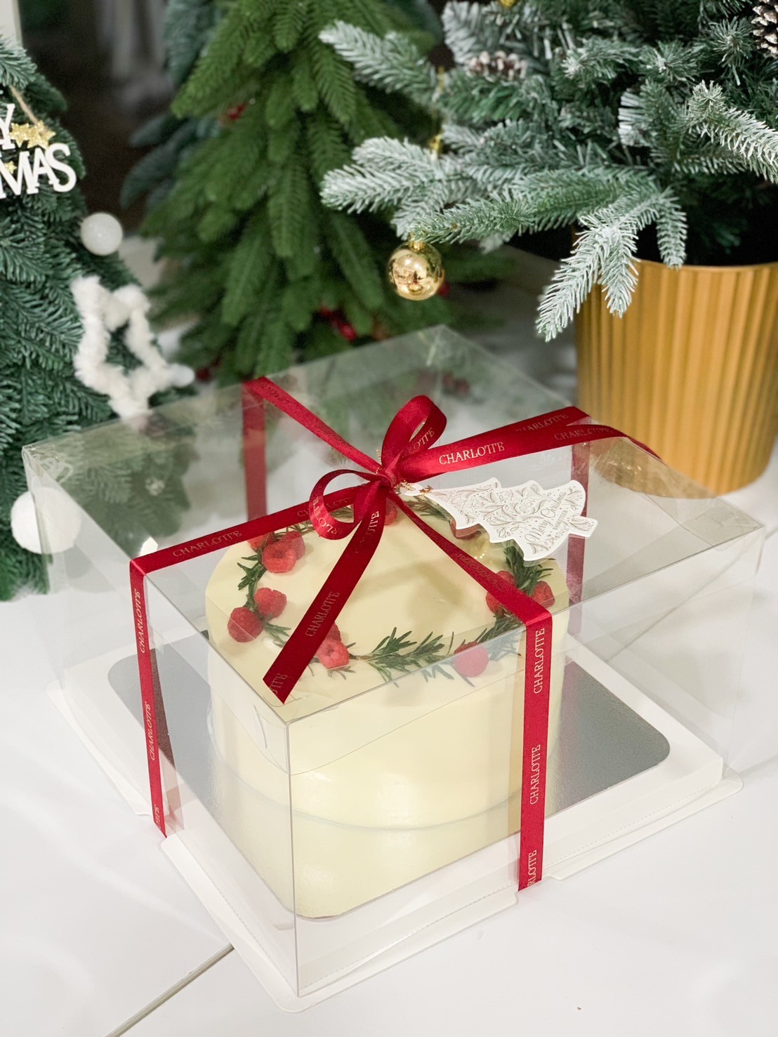 Elegant strawberry Christmas cake in a transparent gift box with red ribbons, surrounded by holiday decorations and Christmas trees.