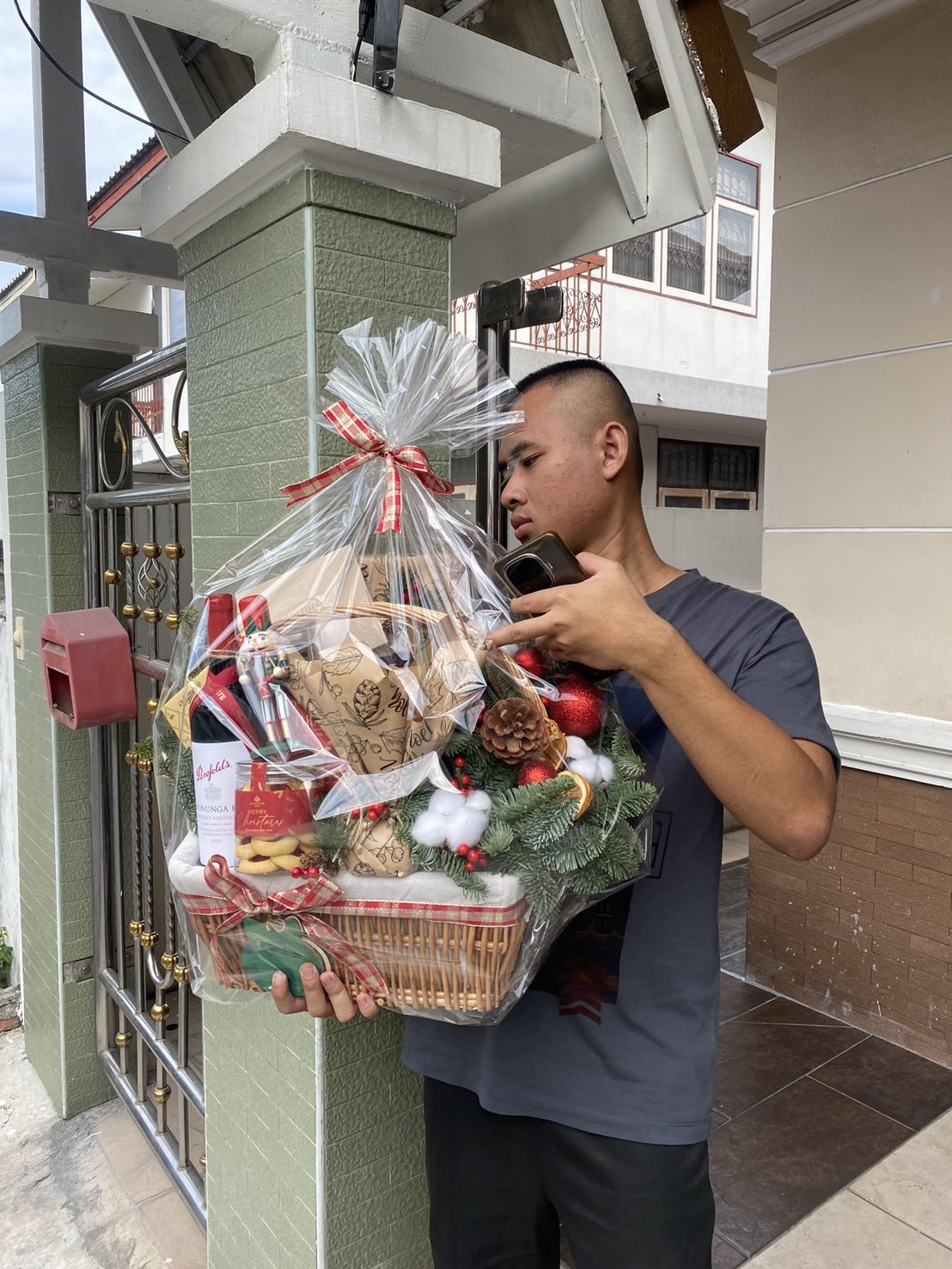 A man holding a large festive gift basket wrapped in clear plastic and tied with a red and white ribbon, filled with holiday treats, decorations, and gourmet items, standing outside a residential home.