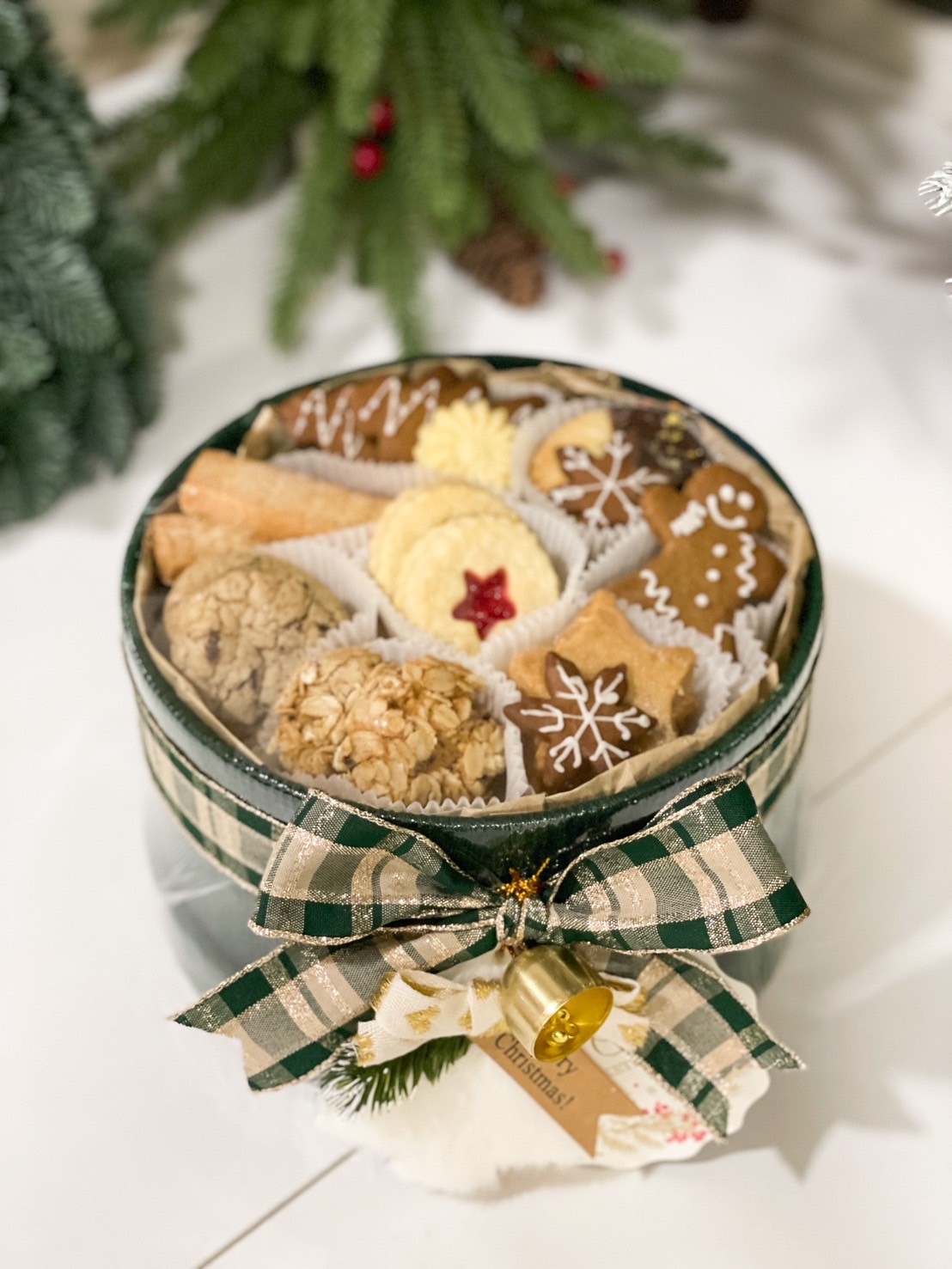 Festive Christmas gift box with assorted cookies, including gingerbread, snowflake designs, and almond treats, decorated with a plaid ribbon and bell.