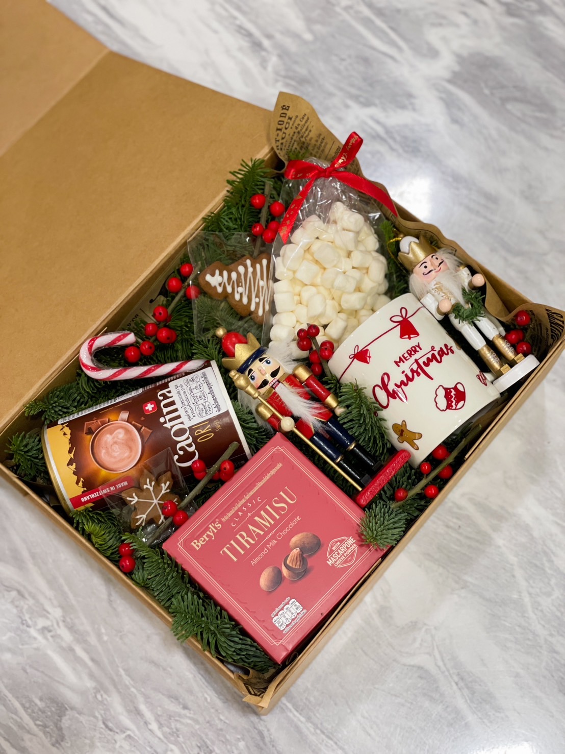 A festive Christmas gift box featuring a nutcracker, tiramisu almonds, butter cookies, a "Merry Christmas" mug, marshmallows, candy canes, and a gingerbread cookie, beautifully arranged with pine greenery, red berries, and holiday decorations, perfect for celebrating the holiday season.