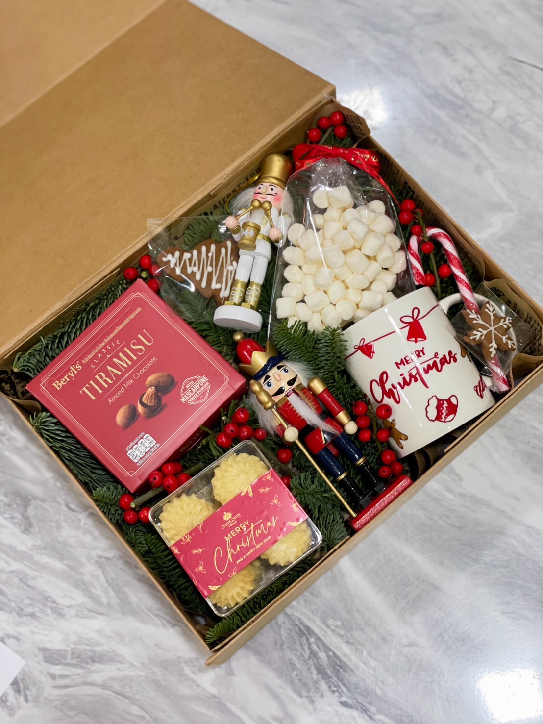 A festive Christmas gift box featuring a nutcracker, tiramisu almonds, butter cookies, a "Merry Christmas" mug, marshmallows, candy canes, and a gingerbread cookie, beautifully arranged with pine greenery, red berries, and holiday decorations, perfect for celebrating the holiday season.