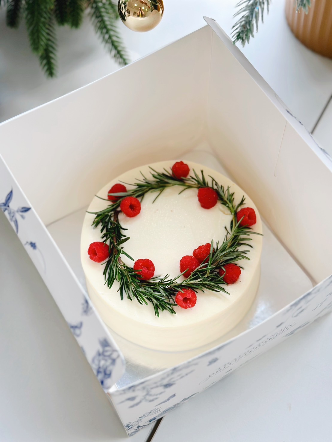 A beautifully decorated Christmas cake in a white box, topped with a festive wreath made of fresh rosemary and vibrant red berries, set beside a Christmas tree adorned with ornaments, perfect for holiday celebrations.