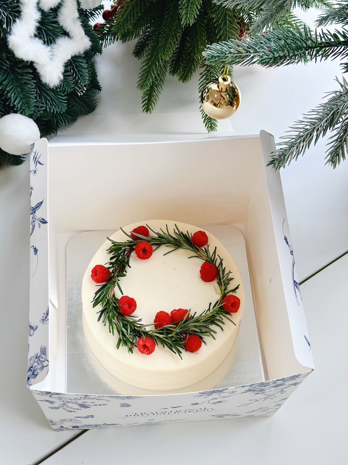 A beautifully decorated Christmas cake in a white box, topped with a festive wreath made of fresh rosemary and vibrant red berries, set beside a Christmas tree adorned with ornaments, perfect for holiday celebrations.