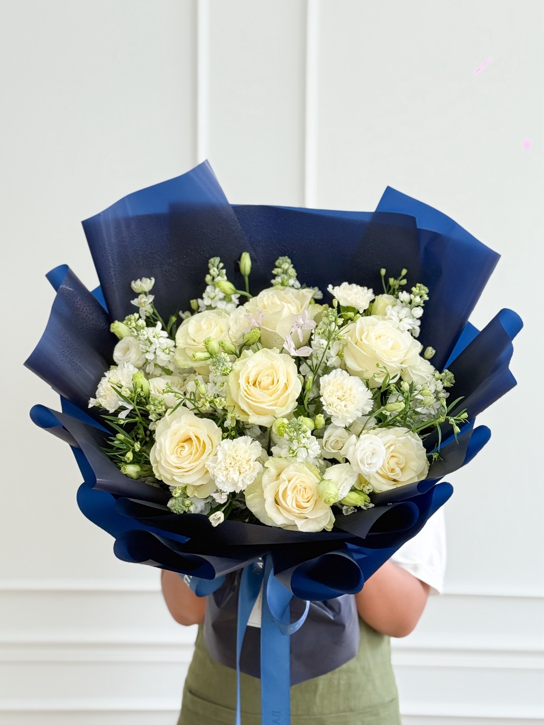 A person in a green apron holds a Graceful White Rose Bouquet and flowers, elegantly wrapped in dark blue paper, against a white background.