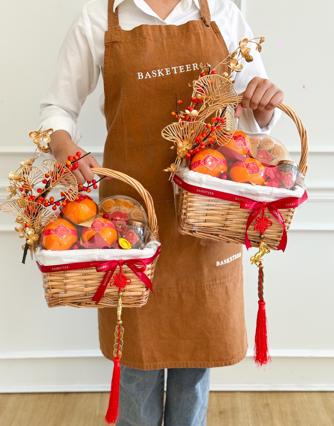 A person wearing a brown apron holds two Festive Harmony Mandarin Baskets filled with oranges and festive items, adorned with red ribbons, tassels, and gold accents.