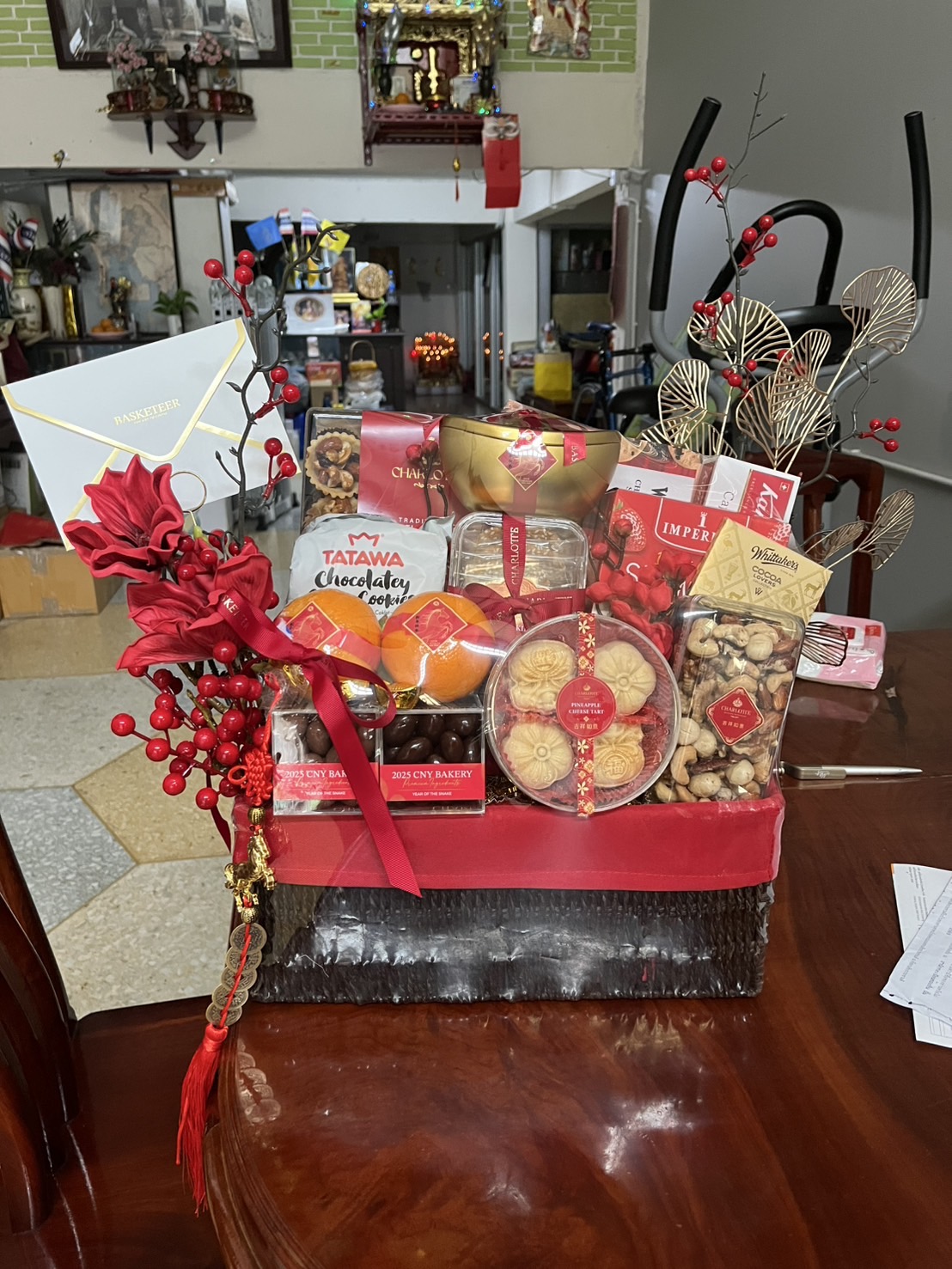 A Prosperity Luxe Chinese New Year Hamper wrapped in red, filled with snacks, biscuits, chocolates, oranges, crisps, and tea, decorated with faux red berries and gold leaves, sits on a wooden table in a living room.