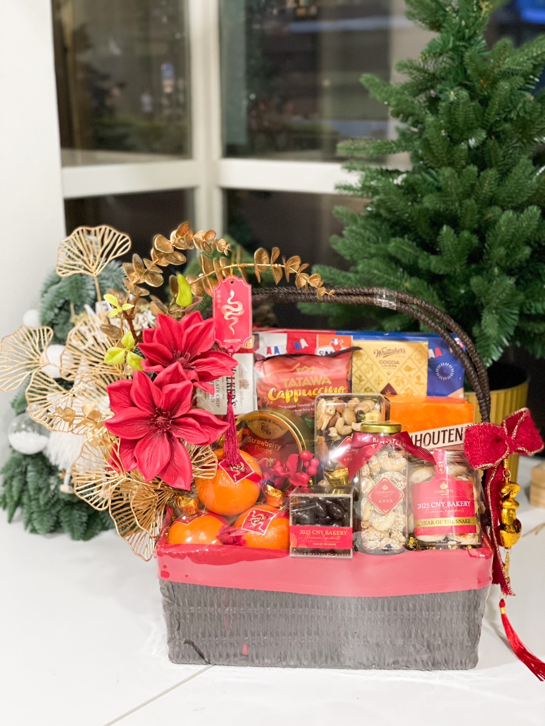 A festive gift basket filled with mandarin oranges, chocolates, cookies, nuts, and premium bakery items, adorned with red and gold floral decorations and New Year embellishments.