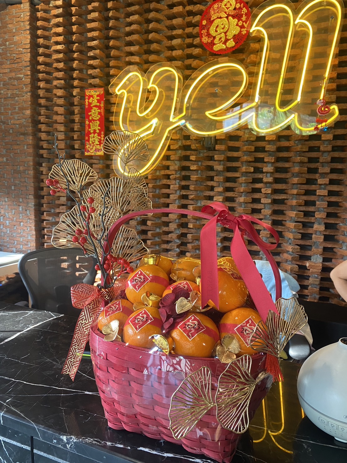 A beautifully decorated Chinese New Year orange basket featuring vibrant oranges, gold accents, red ribbons, and intricate golden ginkgo leaves, placed on a marble countertop with festive red and gold decorations in the background.