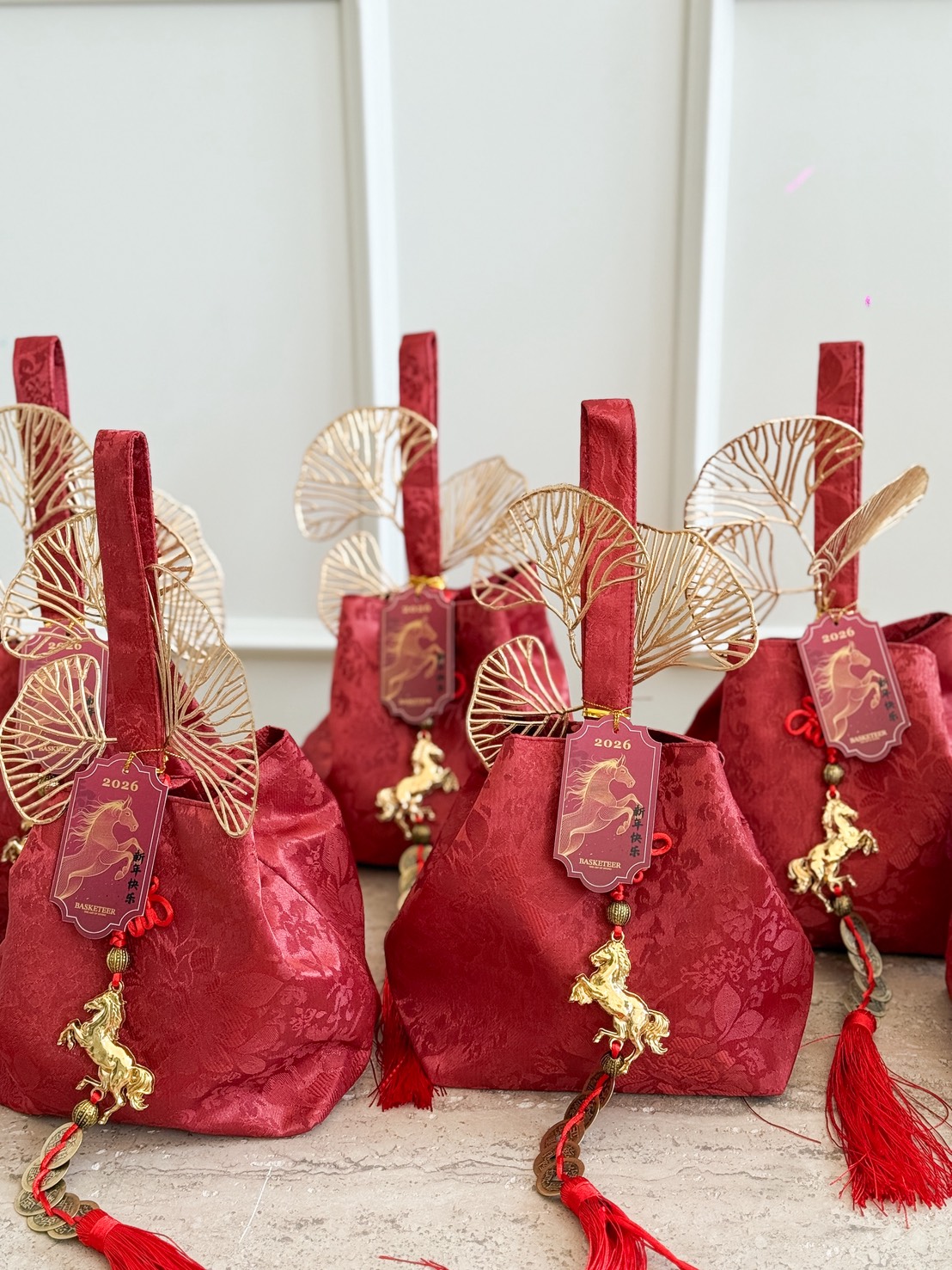 Red Prosperity Mandarin Gift Bags with gold accents, horse charms, and red tassels are arranged on a light surface. Each bag features a gold fan ornament and a red "2026" tag with a horse illustration for festive gifting.