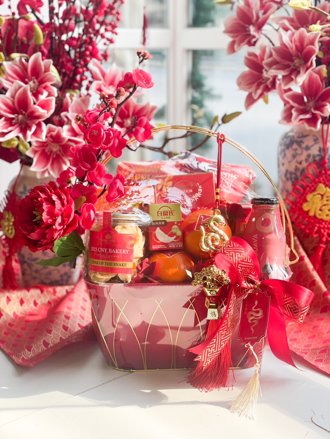 Elegant Chinese New Year hamper featuring fresh mandarin oranges, festive cookies, and prosperity charms in a gold-handled red basket with floral accents.