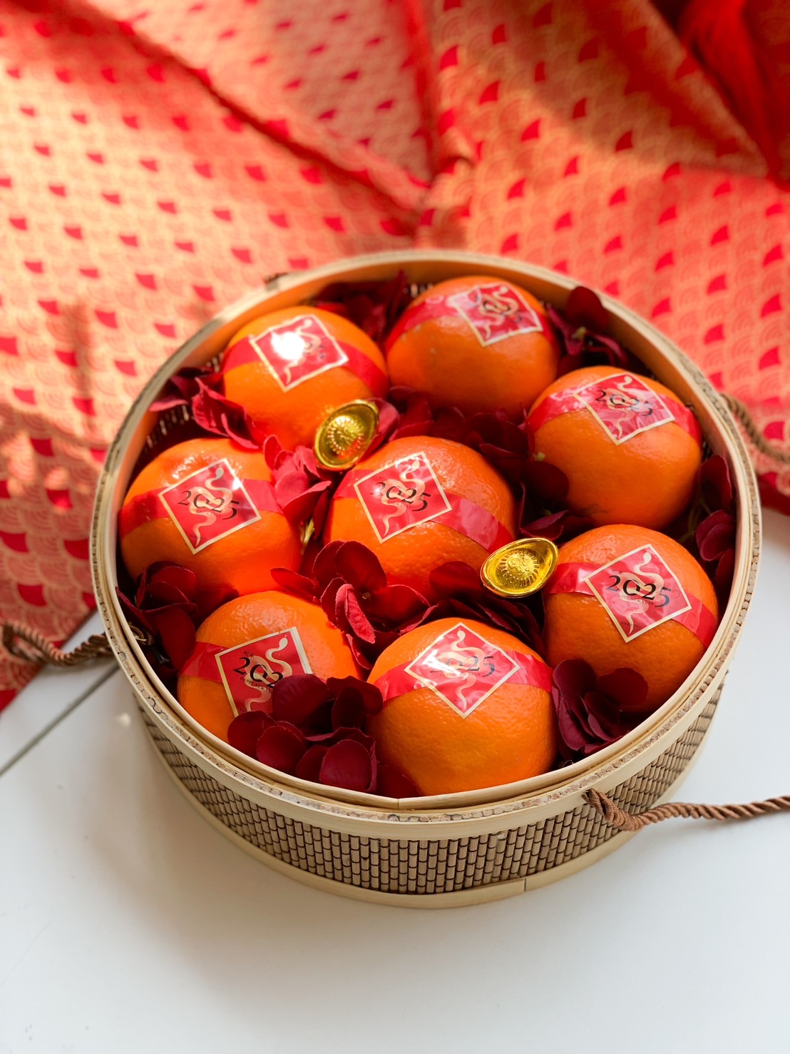 Close-up of a Lunar New Year Mandarin oranges arranged in a circular bamboo box, decorated with red ribbon and festive charm.