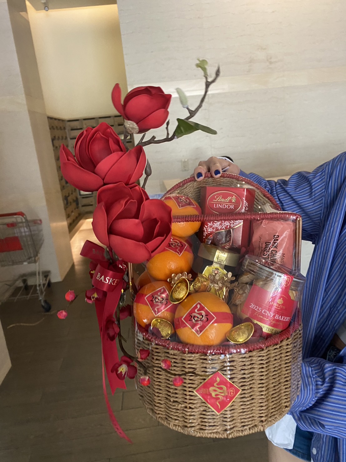 Chinese New Year gift basket with mandarin oranges, chocolates, festive treats, gold ingots, and red floral decorations for prosperity.