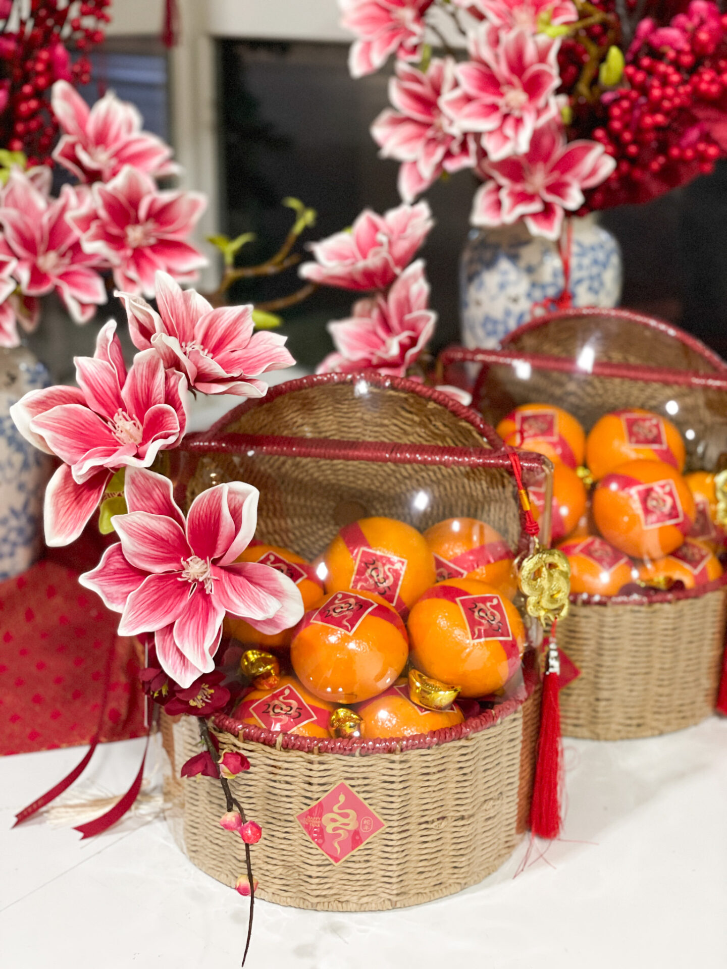 Chinese New Year woven gift basket with mandarin oranges, golden ingots, red floral accents, and a clear dome cover for prosperity.