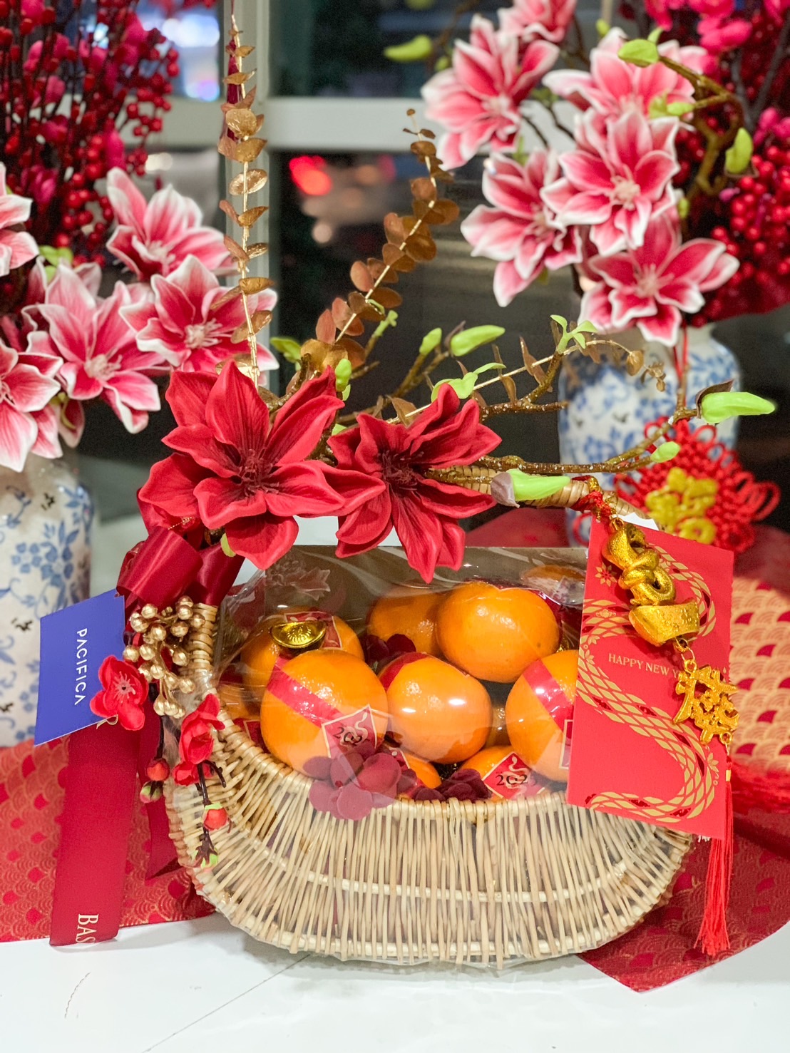 Chinese New Year rattan gift basket with mandarin oranges, gold ingots, red floral accents, and lucky charms for prosperity.