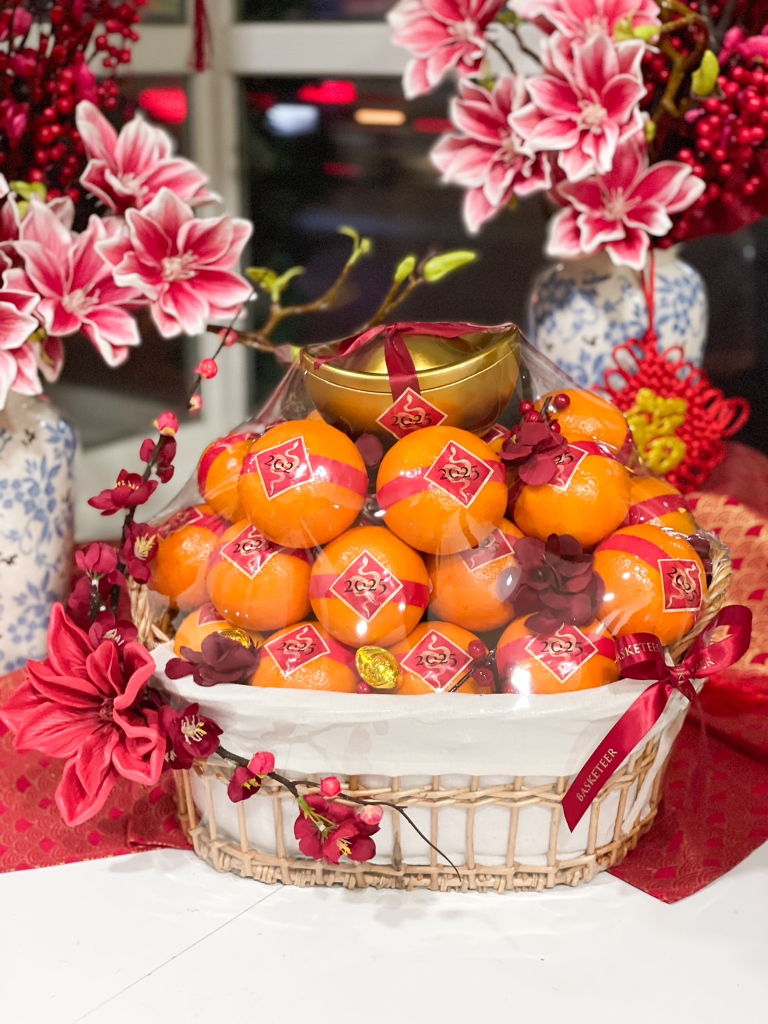 Chinese New Year gift basket with mandarin oranges, gold ingot, red floral decorations, and a clear dome cover for prosperity and wealth.