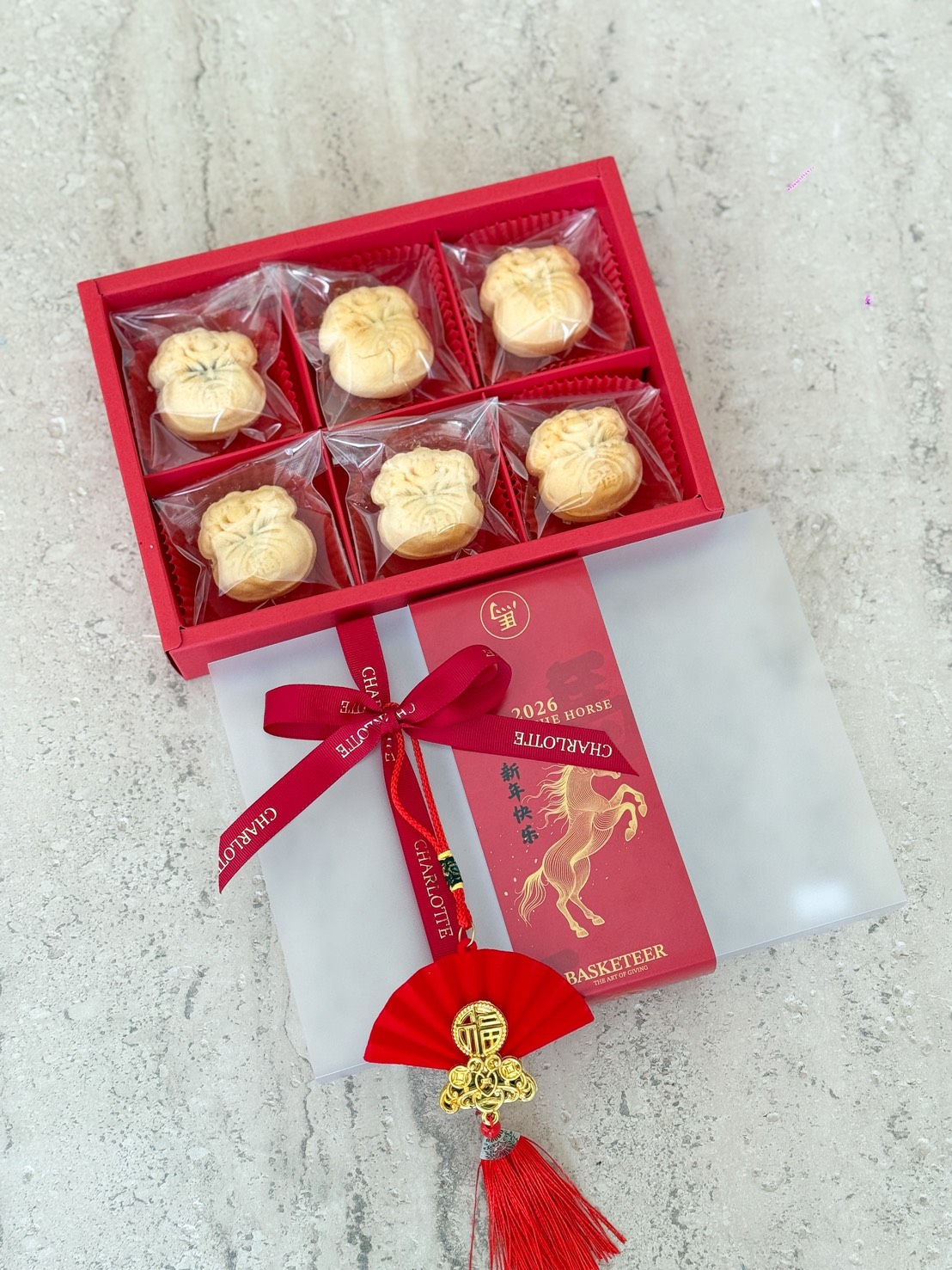 A red box of six orange cheese tart gift pastries wrapped in clear plastic, partially covered by a frosted lid with gold lettering, red ribbon, and decorative tassel featuring a fan and horse design, displayed on a light marble surface.