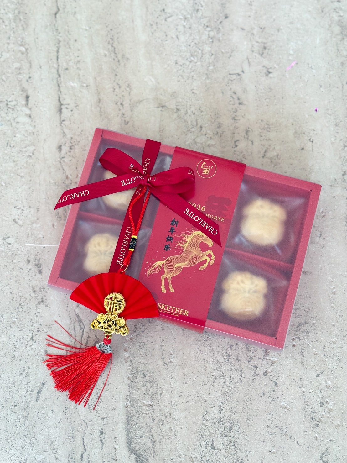 A red gift box with four mooncakes and Orange Cheese Tarts Gift, decorated with a ribbon and a tag featuring a golden horse. A red tassel with a fan-shaped charm and gold accents is attached, resting on a light marble surface.