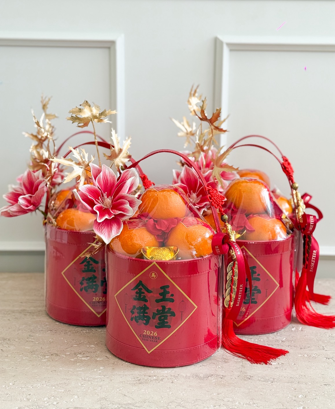 Three red Elegant Prosperity Orange Buckets for Chinese New Year are filled with oranges, gold-wrapped chocolates, artificial red flowers, and golden leaves, adorned with red tassels and sitting on a light surface before a white wall.