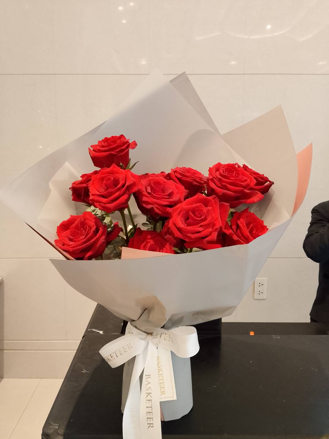 A Velvet Crimson Rose Bouquet, wrapped in white paper and tied with a white ribbon, sits on a black surface against a white tiled background.