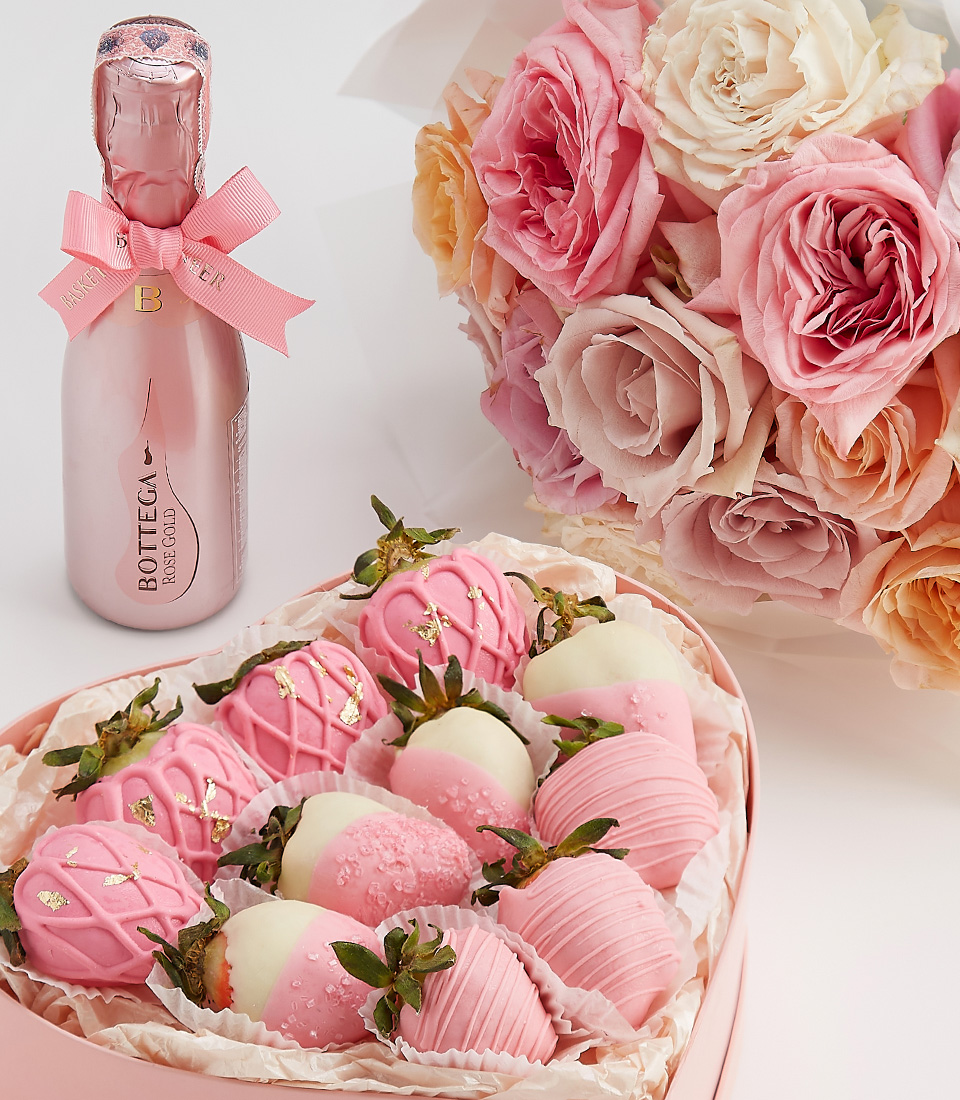 Close-up of a heart-shaped gift box with pink and white chocolate-covered strawberries, gold accents, and a bottle of sparkling rosé.