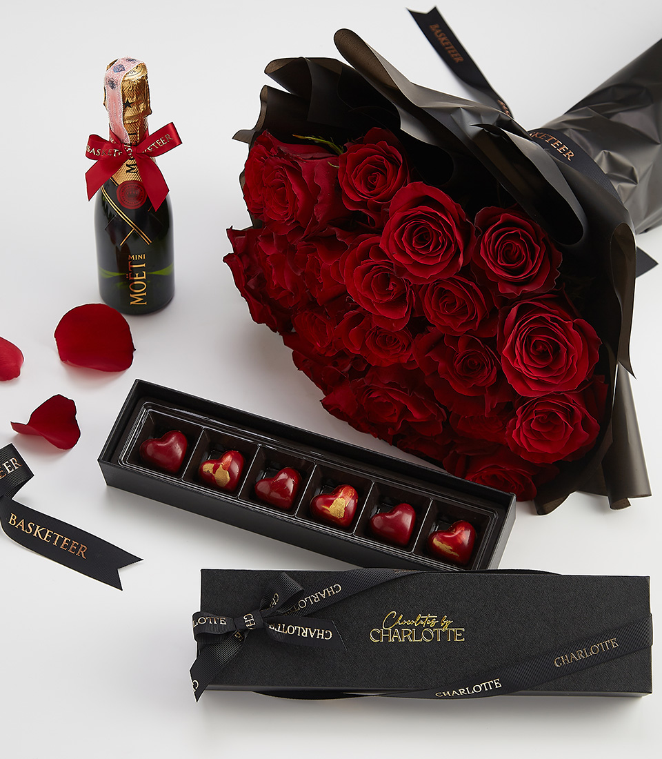 Introducing the Moët & Heart Chocolate Red Rose Gift Set: A bouquet of red roses wrapped in black paper, paired with a bottle of sparkling wine and a box of heart-shaped chocolates, elegantly arranged on a white surface. Scattered rose petals and black ribbons complete this exquisite display.