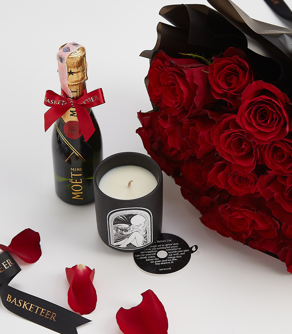 Close-up of a premium romantic gift set including a bouquet of deep red roses, a mini Moët champagne bottle, and a black luxury scented candle, elegantly arranged with rose petals and a black Basketeer ribbon.