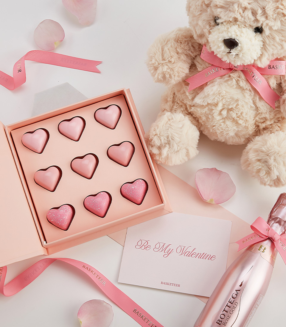A romantic Valentine’s gift with a plush teddy bear, pink heart chocolates, Bottega Rose Gold Champagne, and a heartfelt greeting card.