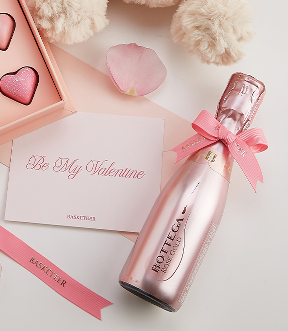 Close-up of a romantic Valentine’s gift with a plush teddy bear, pink heart chocolates, Bottega Rose Gold Champagne, and a heartfelt greeting card.