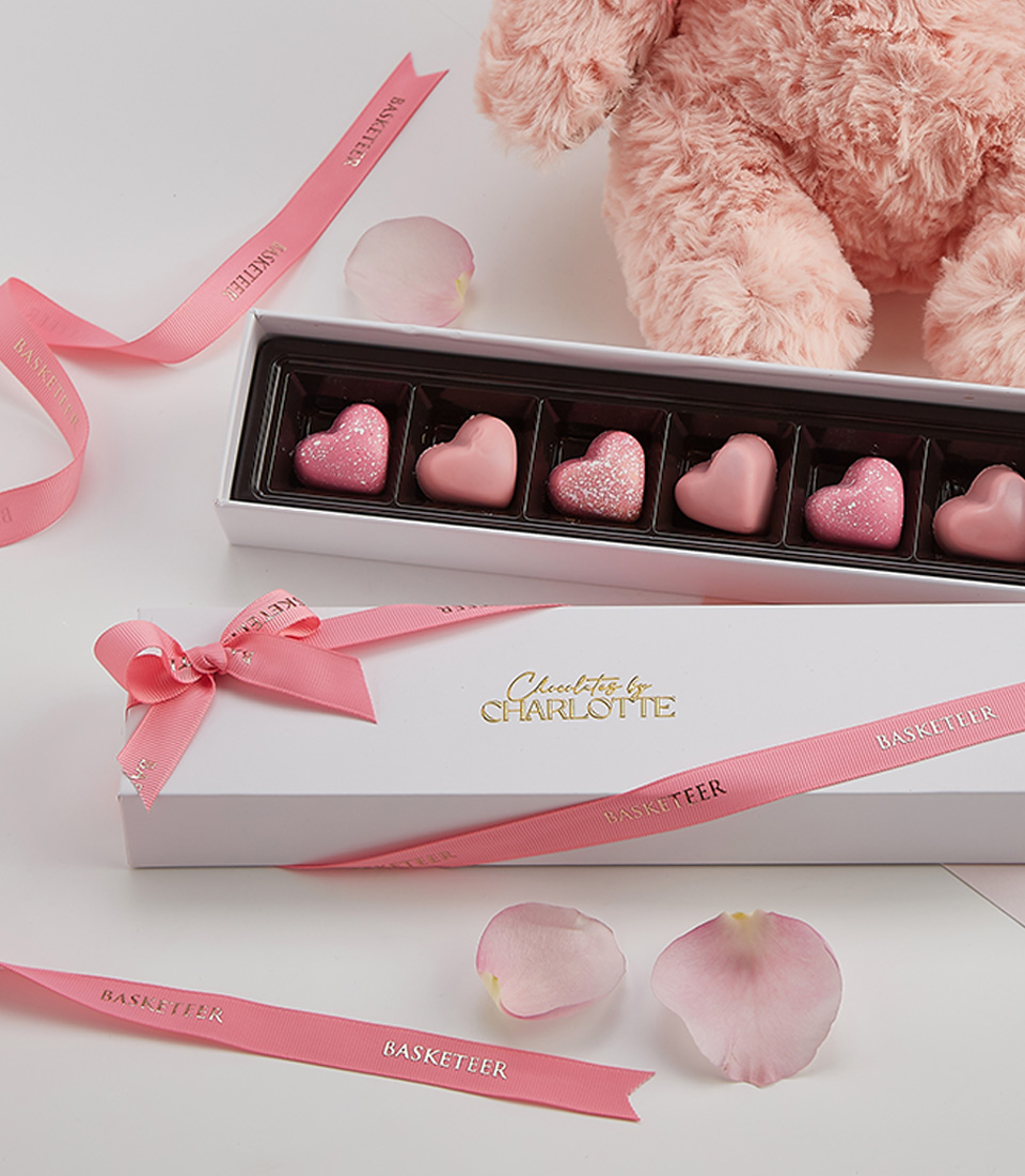Close-up of a charming pink teddy bear with a Basketeer ribbon, paired with a box of heart-shaped chocolates in an elegant white and pink-themed gift set.