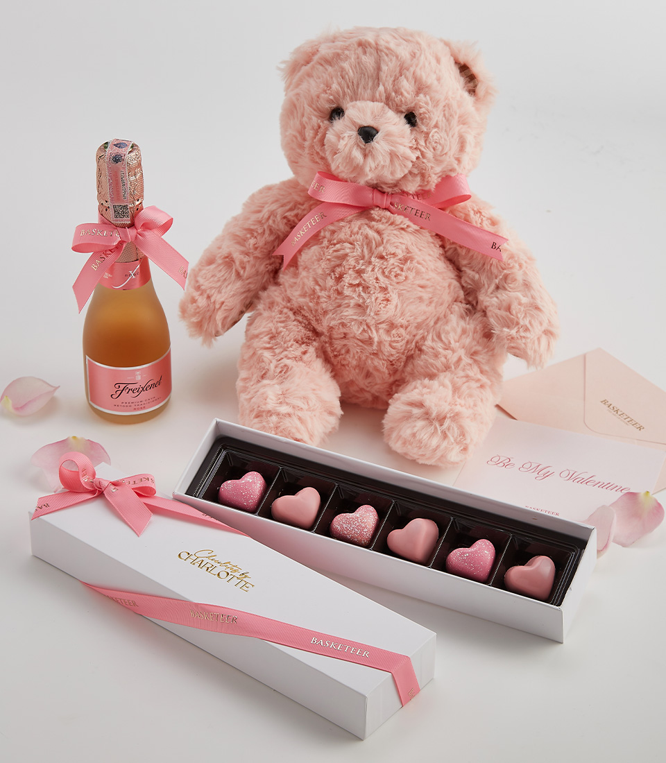 A romantic gift set featuring a blush teddy bear, heart-shaped chocolates, and a bottle of sparkling rosé, presented with a Valentine’s card.