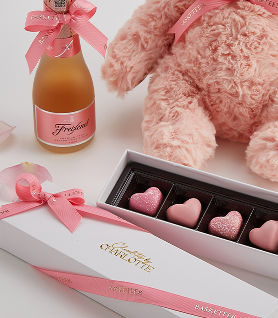 Close-up of a romantic gift set featuring a blush teddy bear, heart-shaped chocolates, and a bottle of sparkling rosé, presented with a Valentine’s card.