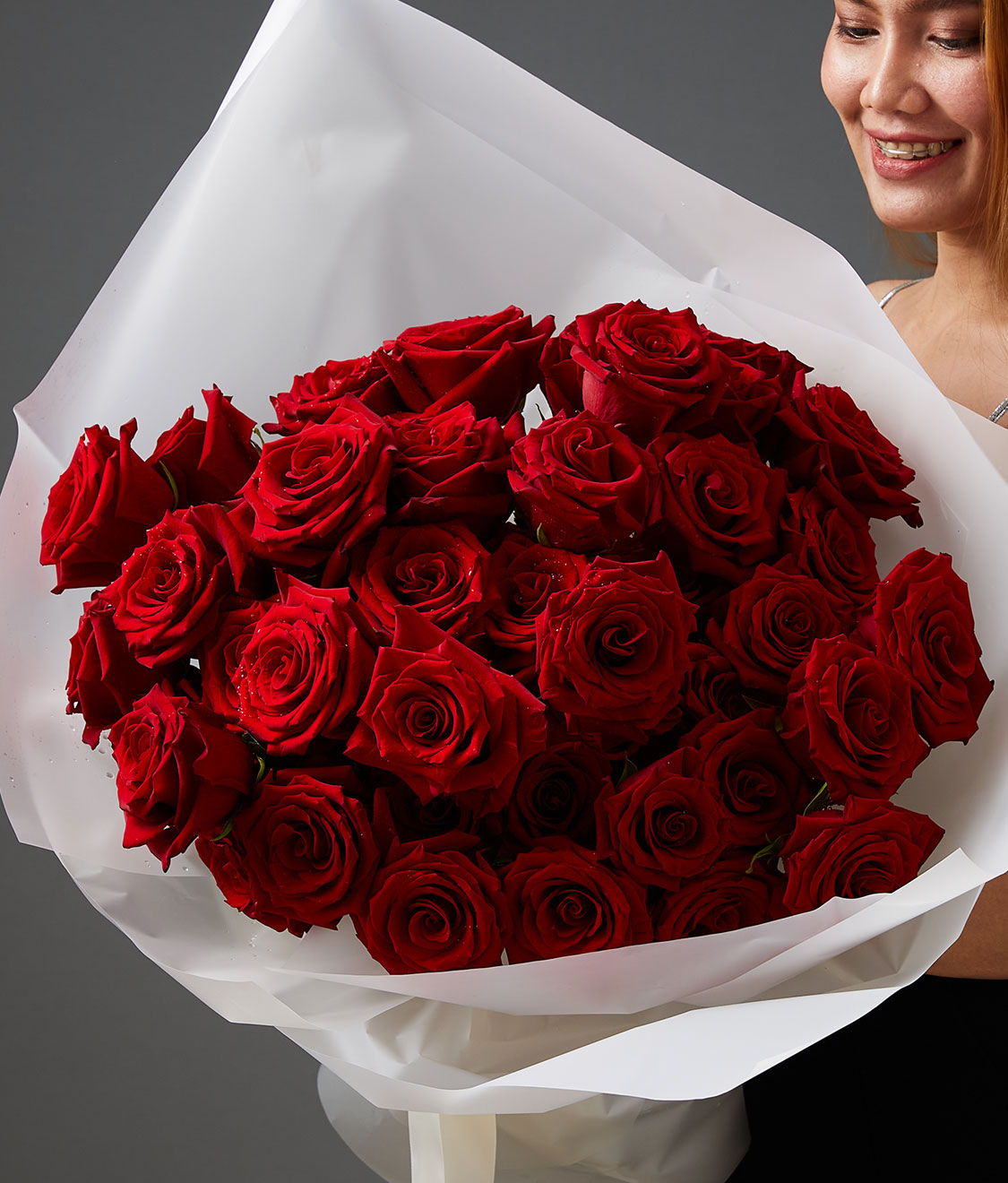 A person holding a large bouquet of vibrant red roses wrapped in white paper, smiling softly. The background is a plain dark gray.
