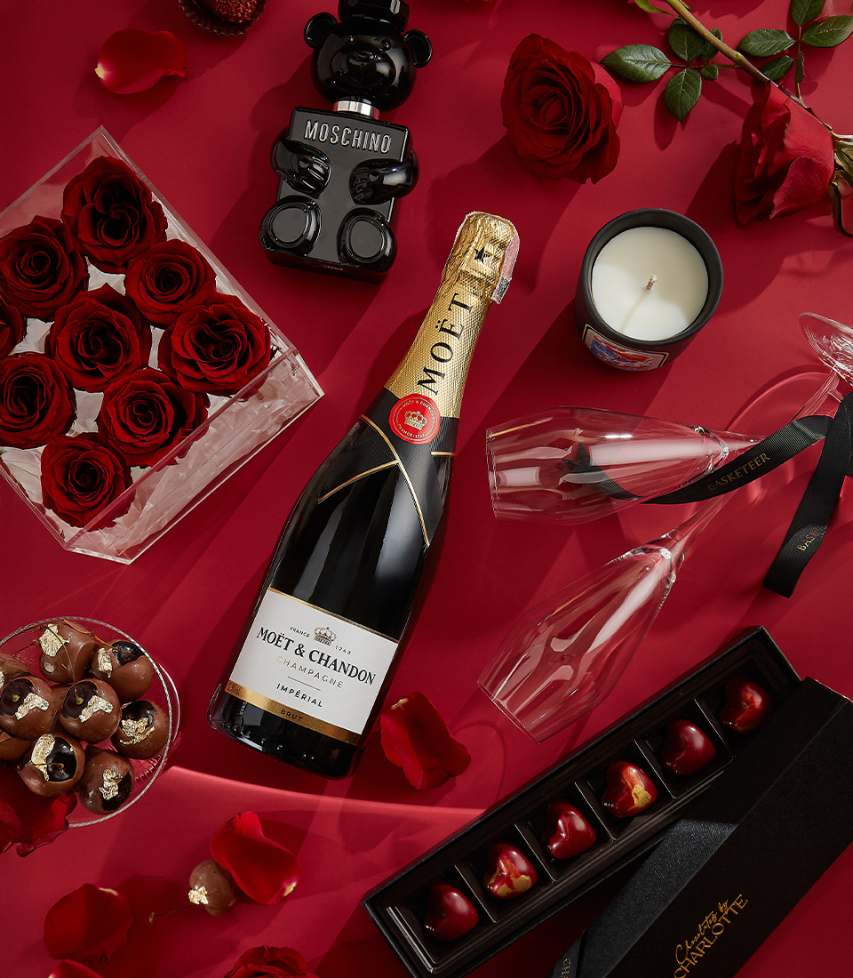 A luxurious Valentine's gift set featuring Moët & Chandon Brut champagne, red preserved roses in an acrylic box, Moschino Toy Boy perfume, gold-accented chocolates, and a scented candle—the ultimate romantic gesture.