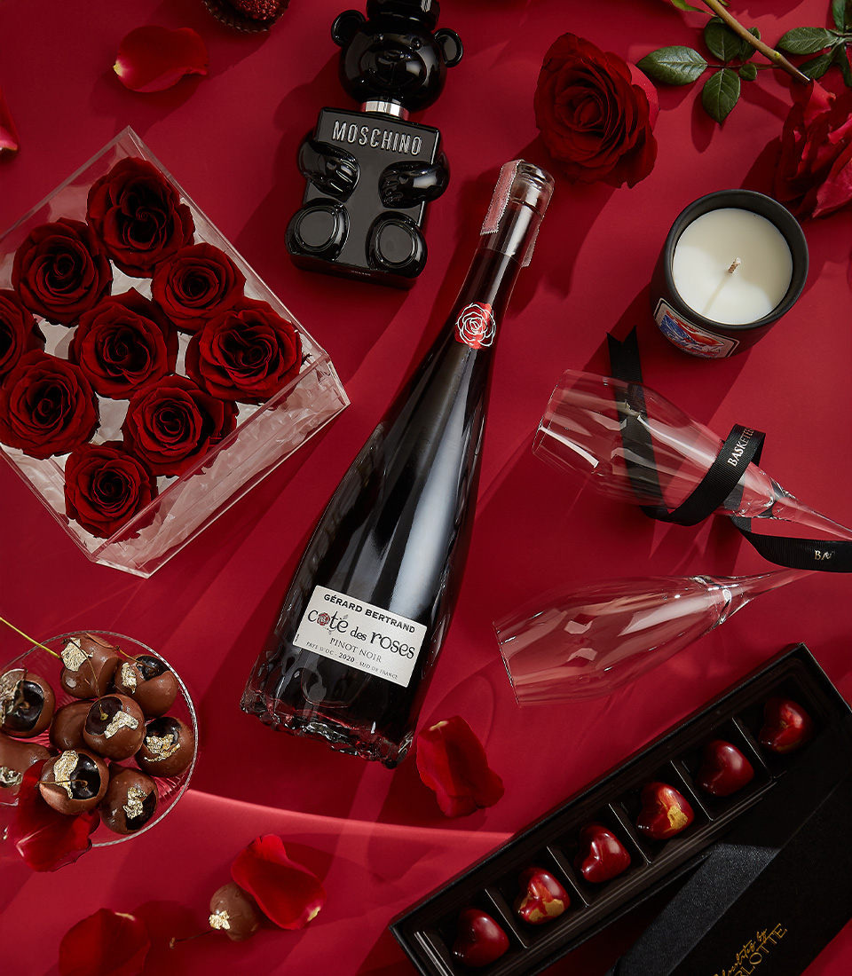 An elegant Valentine's gift set featuring red preserved roses in an acrylic box, Gerard Bertrand Pinot Noir wine, Moschino Toy Boy perfume, heart-shaped chocolates, and a scented candle—perfect for an unforgettable romantic gesture.