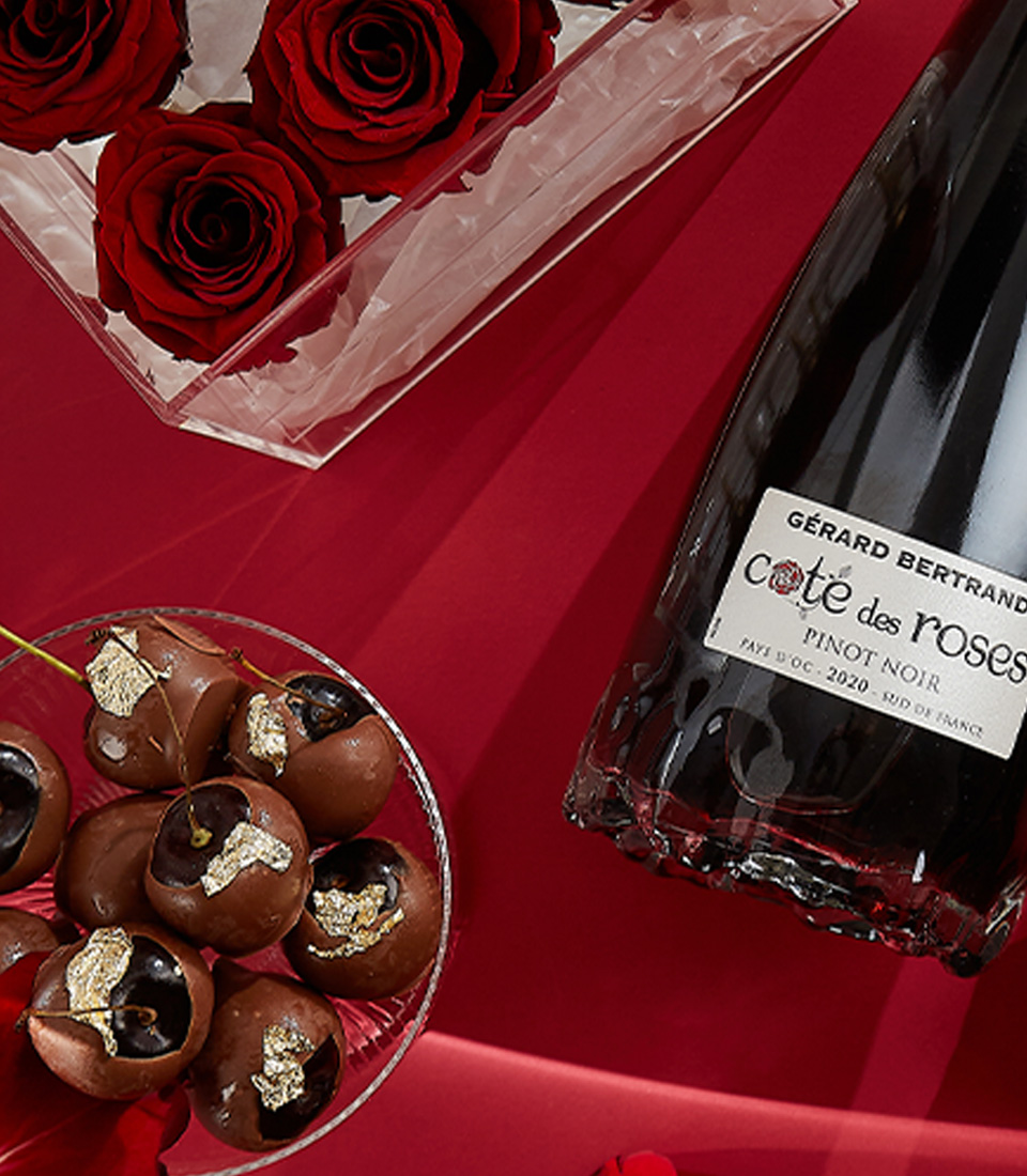 Close-up of a elegant Valentine's gift set featuring red preserved roses in an acrylic box, Gerard Bertrand Pinot Noir wine, Moschino Toy Boy perfume, heart-shaped chocolates, and a scented candle—perfect for an unforgettable romantic gesture.
