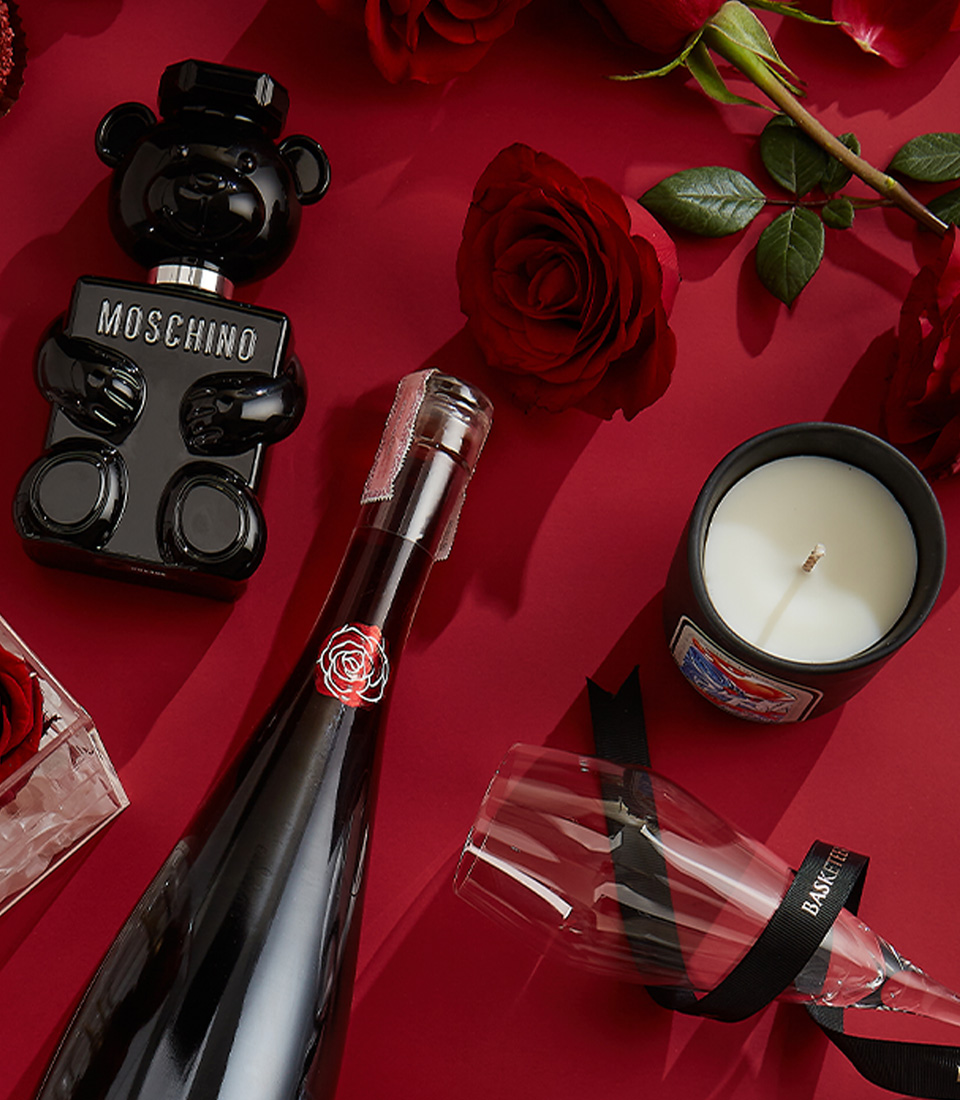 Close-up of a elegant Valentine's gift set featuring red preserved roses in an acrylic box, Gerard Bertrand Pinot Noir wine, Moschino Toy Boy perfume, heart-shaped chocolates, and a scented candle—perfect for an unforgettable romantic gesture.