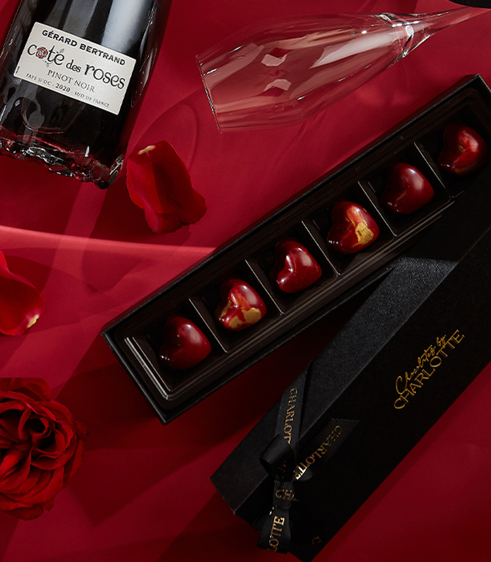 Close-up of a elegant Valentine's gift set featuring red preserved roses in an acrylic box, Gerard Bertrand Pinot Noir wine, Moschino Toy Boy perfume, heart-shaped chocolates, and a scented candle—perfect for an unforgettable romantic gesture.
