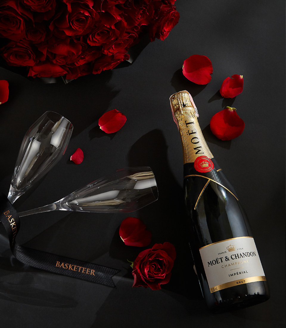 Moët & Chandon champagne bottle with two crystal flutes, surrounded by a bouquet of deep red roses and scattered rose petals on a black background with a Basketeer ribbon.