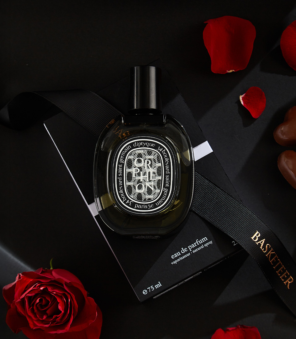 Close-up of a Luxury gift set featuring a bouquet of deep red roses, a designer perfume bottle, and a box of heart-shaped chocolates, beautifully arranged on a black background with scattered rose petals.