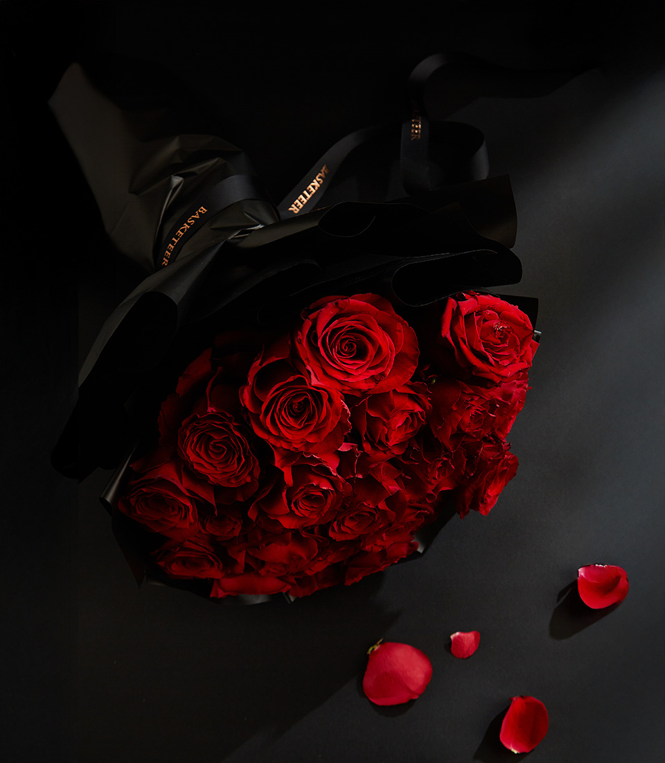 Elegant bouquet of deep red roses wrapped in black paper with a Basketeer ribbon, set against a dark background with scattered rose petals.