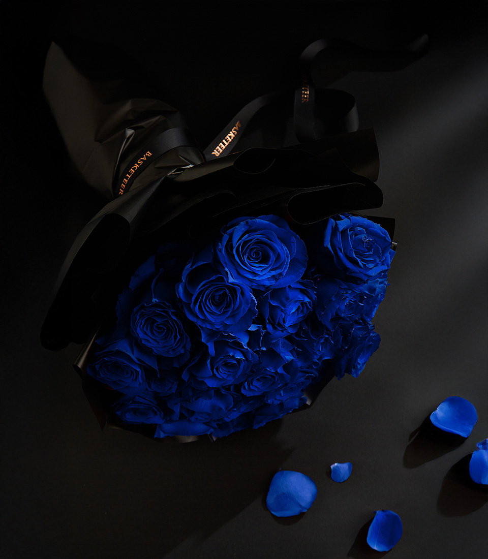 A luxurious bouquet of deep midnight blue roses, wrapped in black paper with a Basketeer ribbon, set against a dark background with scattered blue rose petals.