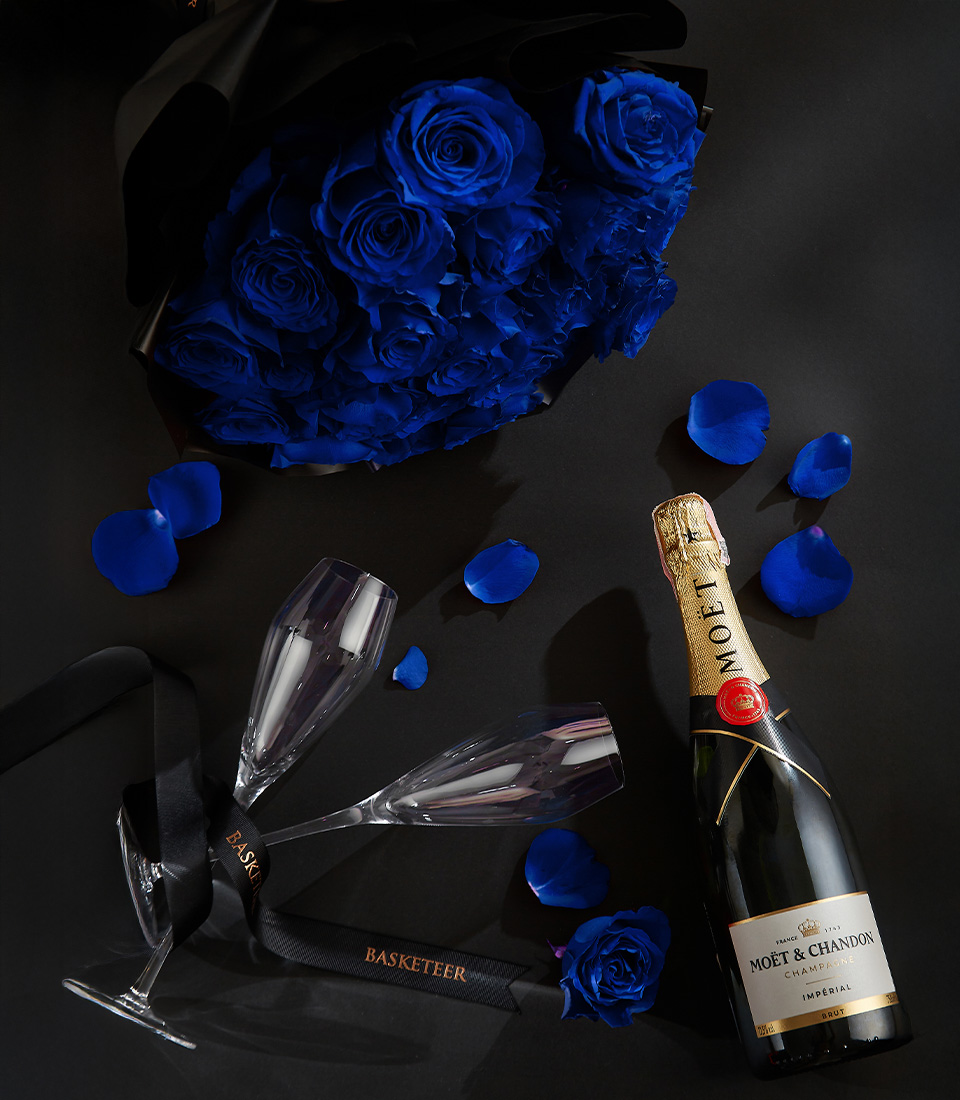 Moët & Chandon champagne bottle with two crystal flutes, surrounded by royal blue roses and petals on a black background, with a Basketeer ribbon.