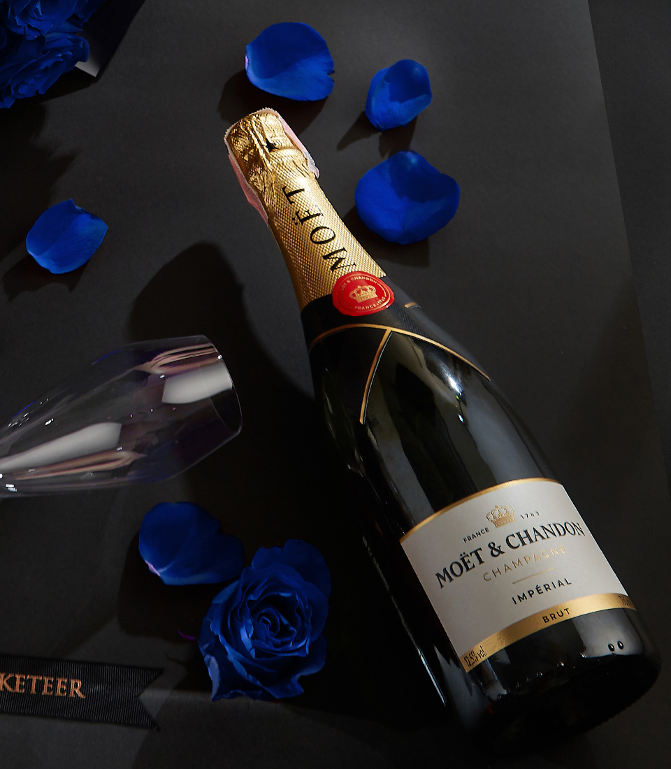 Moët & Chandon champagne bottle with two crystal flutes, surrounded by royal blue roses and petals on a black background, with a Basketeer ribbon.