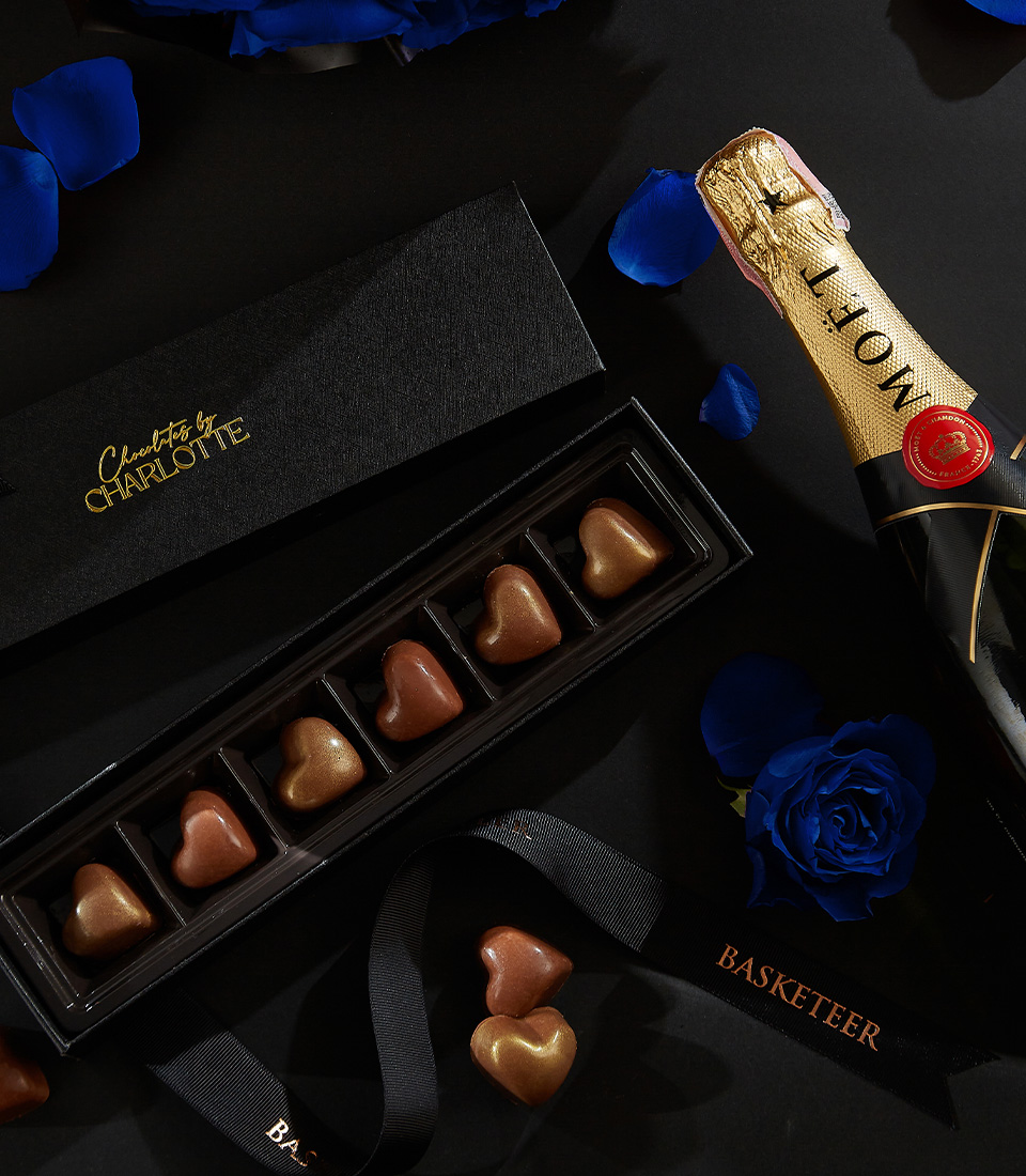Moët & Chandon champagne bottle with a black box of heart-shaped chocolates, deep blue roses, and rose petals on a black background, with a Basketeer ribbon.
