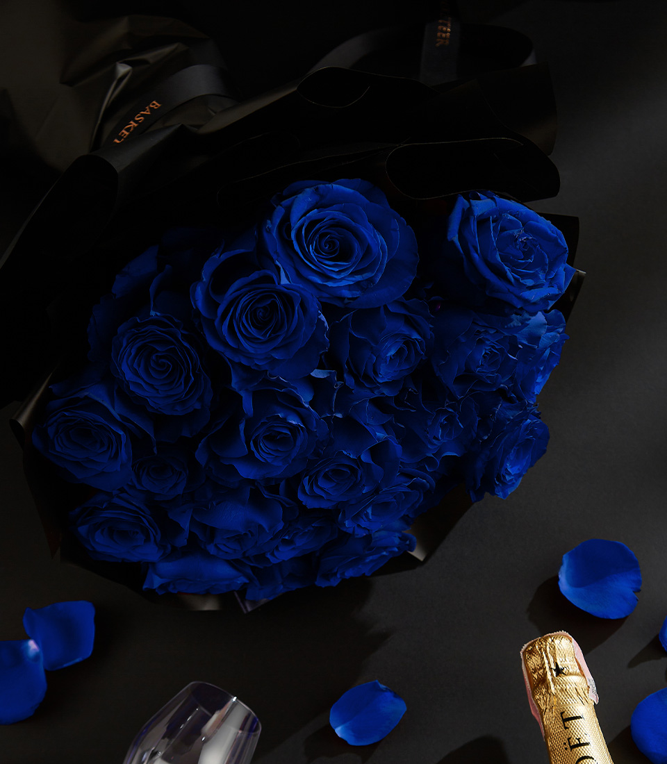 Moët & Chandon champagne bottle with a black box of heart-shaped chocolates, deep blue roses, and rose petals on a black background, with a Basketeer ribbon.
